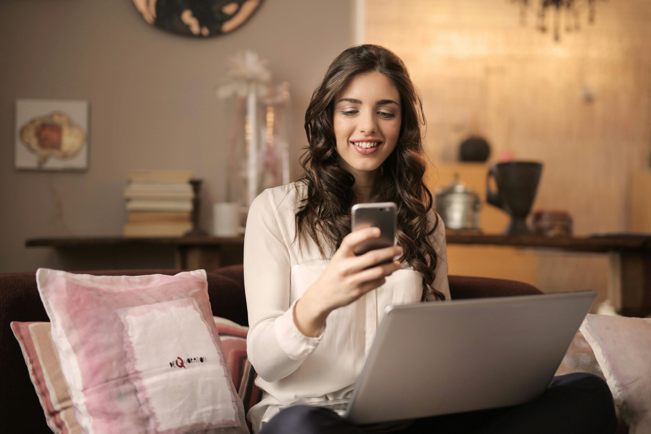 woman sitting on couch holding cell phone with computer on here lap