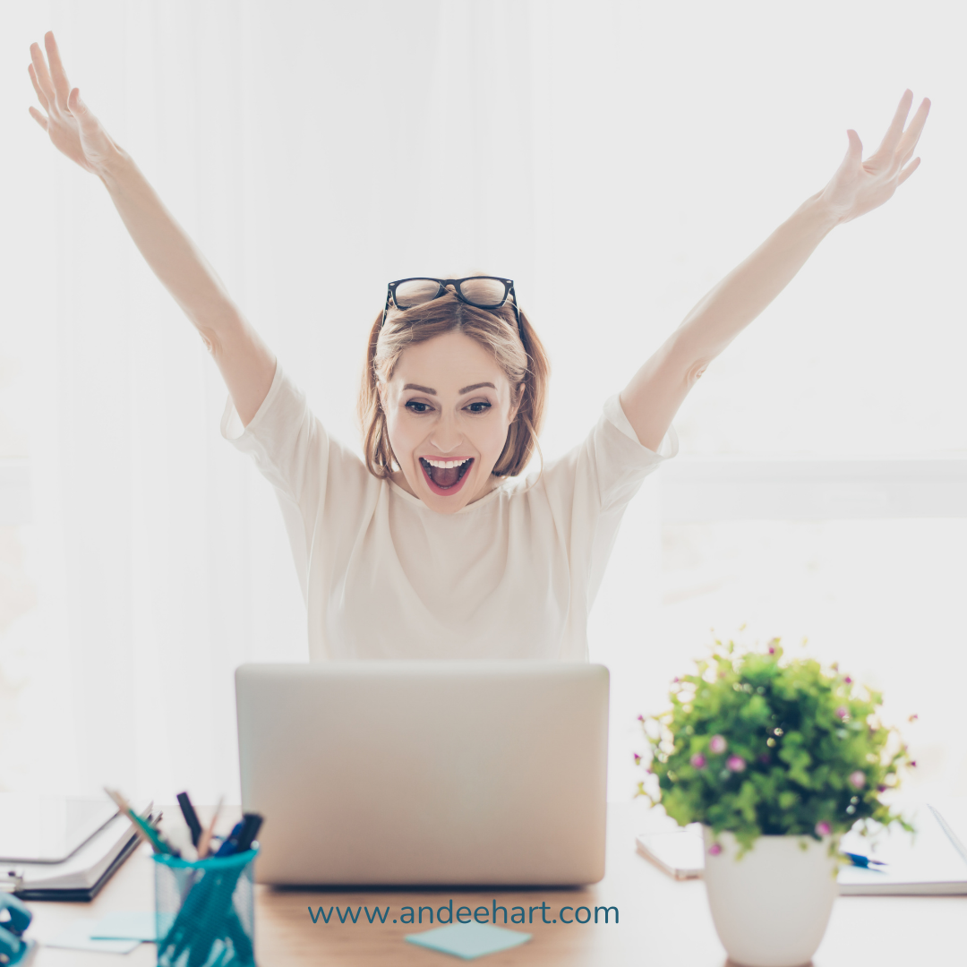 Image is of a female that is throwing her hands in the air in excitement and looking at her computer