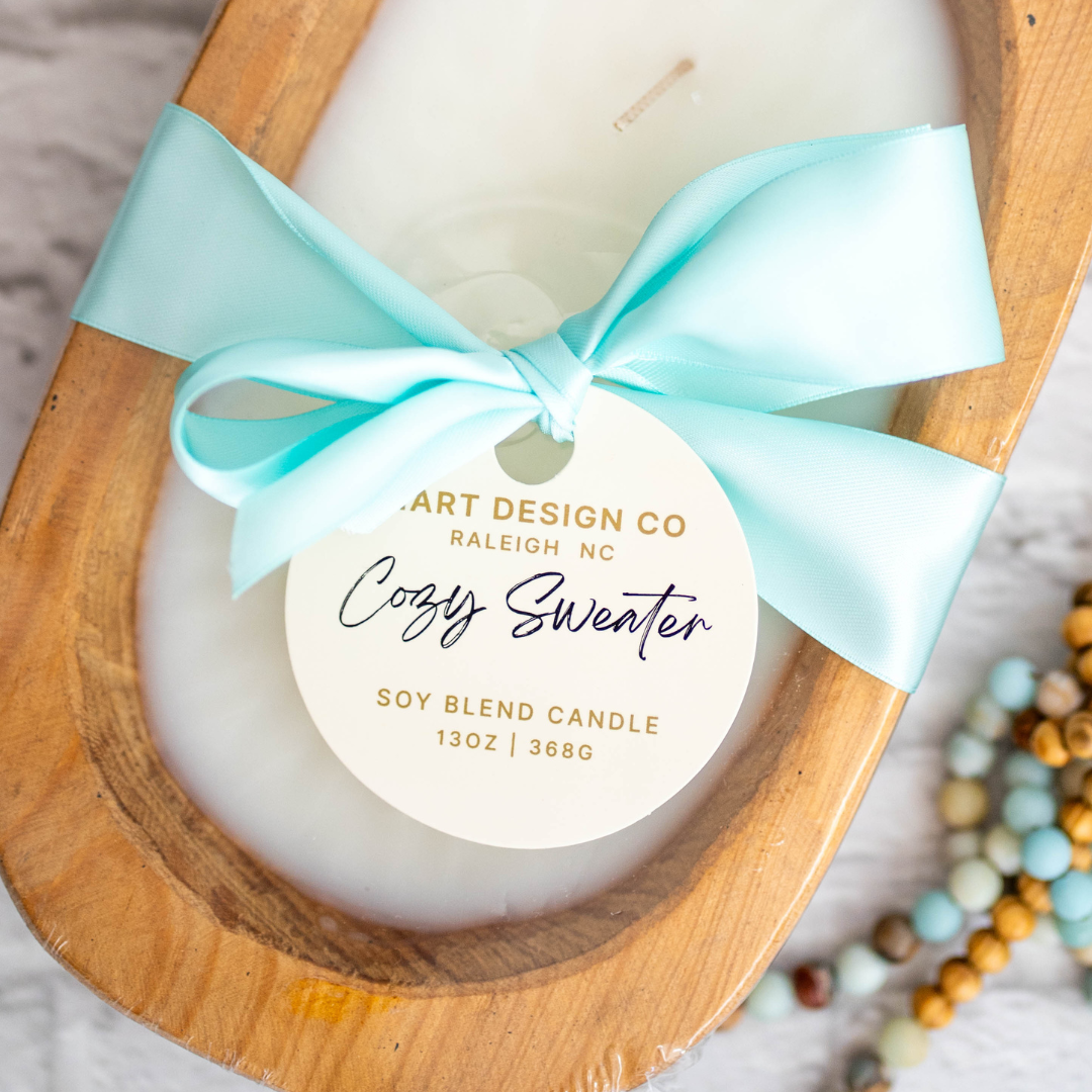 Image is a beautiful soy blend candle from Hart Design co. The candle is in a gorgeous wooden vessel, the candle is white and there is a baby blue bow around the vessel. And the candle scent is cozy sweater.