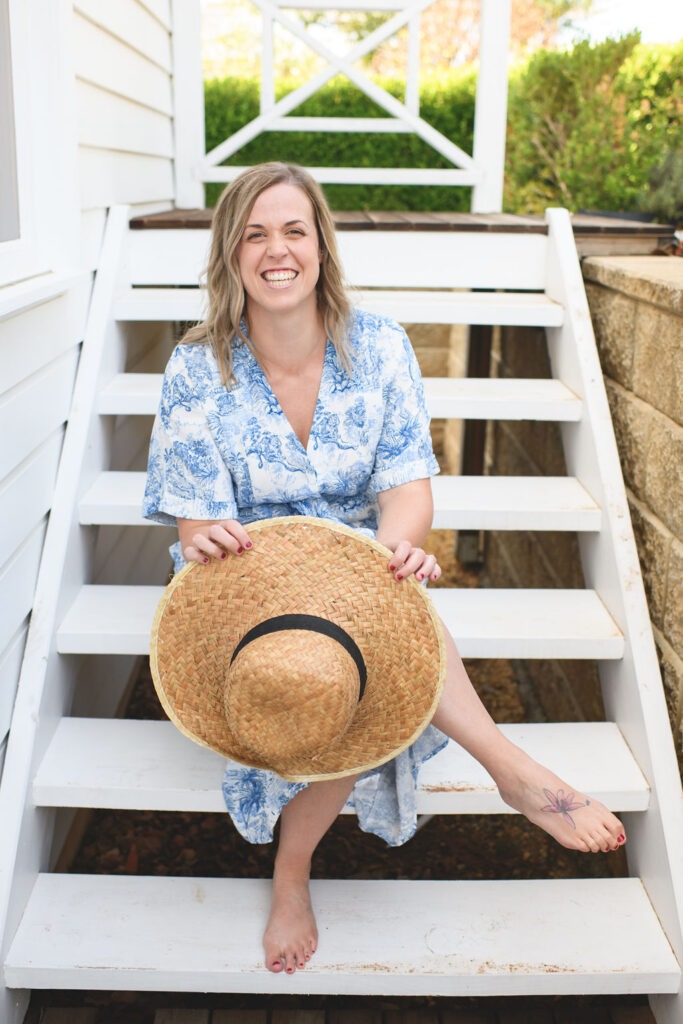 SJ Meeson who is a guest on the She Sells Differently Podcast and she is smiling at the camera while sitting on a set of stairs and holding a big straw hat in her hands.