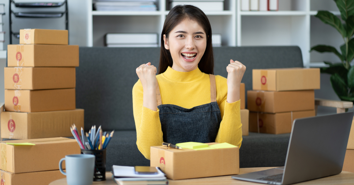 Image is of person sitting at a desk looking excited with their arms in the air and boxes around them