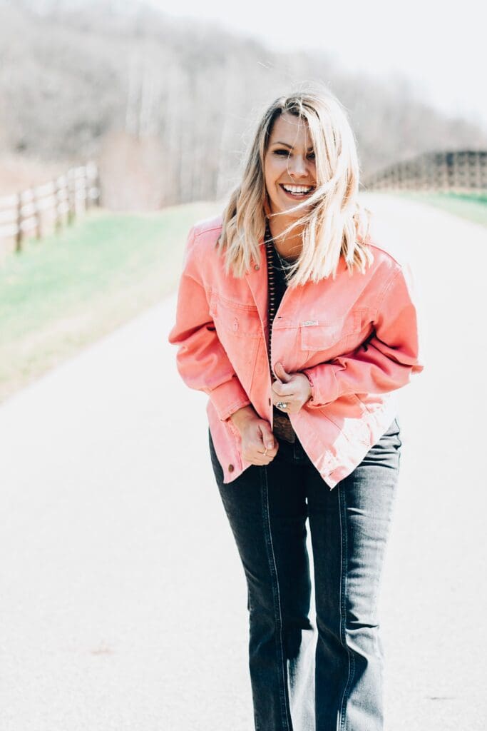 Image is of Ashley Alderson, Founder and CEO of the Boutique hub. She is smiling at the camera in a coral pink jacket and has on black jeans